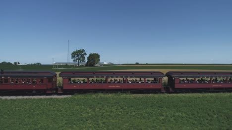 Vista-Aérea-De-Un-Motor-De-Tanque-De-Thomas-Con-Automóviles-De-Pasajeros-Resoplando-A-Lo-Largo-Del-Campo-Amish