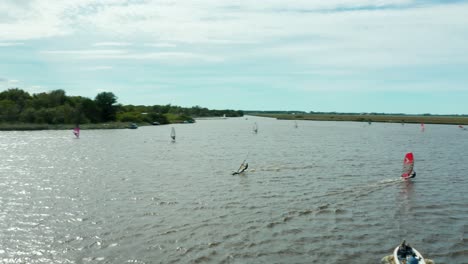 Freizeitsurfer,-Die-Schnell-Mit-Ihrer-Windsurfausrüstung,-Segeln-Und-Brett-über-Den-See-Fahren,-Mit-Schönem-Wind-An-Einem-Sonnigen-Tag,-Drohnen-Parallaxenschuss-4k