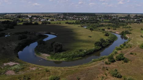 Vista-Aérea-Del-Arroyo-Rodeado-De-árboles-Y-Campos-Con-El-Pueblo-Al-Fondo