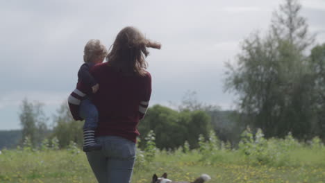 Zeitlupenaufnahme-Einer-Sonnenbeschienenen-Mutter-Und-Ihres-Kindes,-Die-Mit-Ihrem-Hund-Auf-Der-Wiese-Spazieren-Gehen