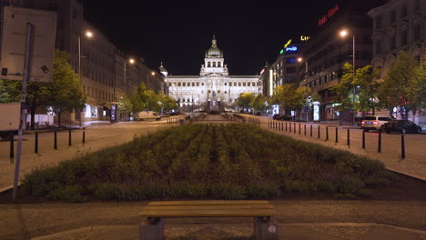 Una-Vista-Del-Museo-Nacional-En-Praga,-Chequia-En-La-Plaza-Vacía-De-Wenceslao-En-El-Centro-Histórico,-Iluminada-Por-Farolas-Durante-Un-Cierre-De-Covid-19,-Pedestal-Disparado-Sobre-Un-Banco-Y-Un-Parche-De-Flores