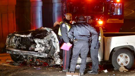 Drei-Polizisten-Unterhalten-Sich-Vor-Einem-Autowrack,-Während-Im-Hintergrund-Ein-Feuerwehrmann-In-Das-Löschfahrzeug-Steigt