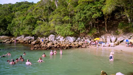 Pan-A-La-Derecha-De-La-Gente-Divirtiéndose-En-El-Mar-Cerca-De-Una-Costa-Rocosa-Y-Bosques-En-La-Playa-Praia-Da-Sepultura,-Brasil