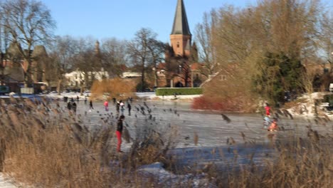 Panorámica-Izquierda-Que-Muestra-A-Personas-Patinando-Sobre-Hielo-En-El-Canal-Con-Caña-En-Primer-Plano-Que-Revela-El-Histórico-Walburgiskerk-Medieval-Y-Drogenapstoren-Levantándose-En-El-Fondo