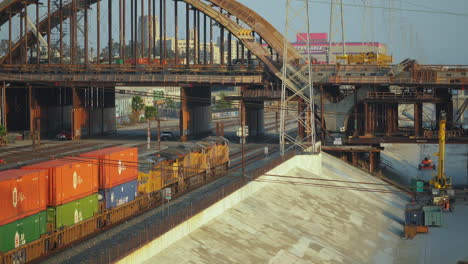 Shot-of-a-container-freight-train-going-slowly-under-the-6th-St-Bridge-in-Downtown-Los-Angeles,-USA-as-sunset