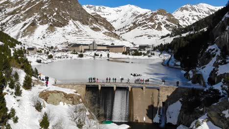 Antena:-Presa-En-Un-Lago-Congelado-Y-Gente-Caminando,-Entre-Montañas-Y-Un-Sharine-En-El-Fondo