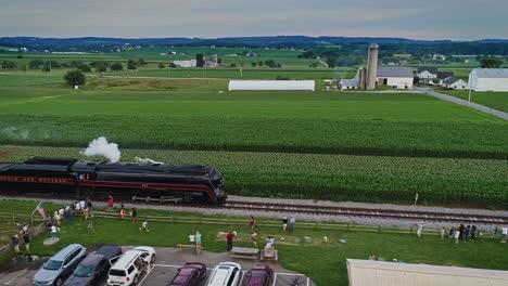 Una-Vista-Aérea-Paralela-De-Un-Antiguo-Tren-De-Pasajeros-A-Vapor-Que-Sopla-Humo-Pasando-Por-Familias-Y-Niños-Observando-Mientras-Viaja-A-Través-De-Las-Tierras-Agrícolas-De-Pensilvania