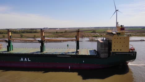 Toma-De-Seguimiento-Aéreo-De-La-Vista-Lateral-Del-Puerto-Del-Buque-De-Carga-Aal-Paris-A-Lo-Largo-De-Oude-Maas-Con-Turbina-Eólica-Inmóvil-En-El-Fondo