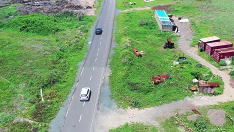 aerial-shot-of-dark-and-white-car-followed-on-a-concrete-road-by-the-coast-with-shore,-foamy-white-waves,-cliff,-buildings-to-right,-cars-and-people-in-the-end,-greenery,-bright-outdoors,-sunshine