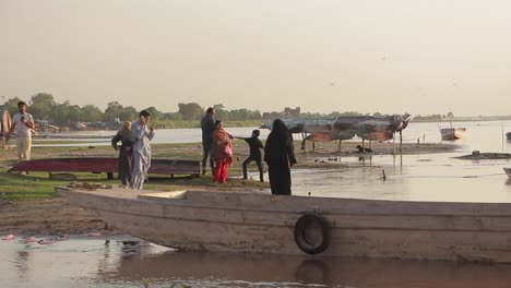 People-Standing-By-A-Boat-On-The-Shore-Of-The-River-Ravi,-Slums-In-Background