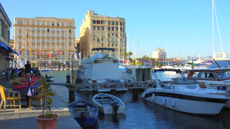 Pan-shot-an-outdoor-cafe-with-tourists-beside-motor-boats-docked-along-the-dock-in-Porto-di-Santa-Lucia-seaport-in-sunny-day-in-Naples,-Italy