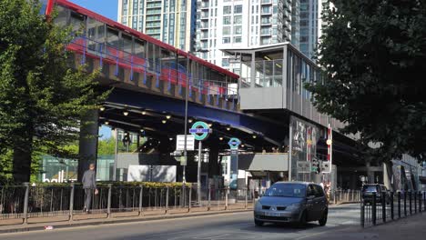 Canary-Wharf,-London,-Vereinigtes-Königreich-–-August-2022-–-Der-DLR-Bahnhof-South-Quay-Mit-Einem-Zug-Auf-Der-Brücke-Darüber-Verlässt-Den-Bahnhof-Und-Unten-Fahren-An-Einem-Sonnigen-Morgen-Autos-Auf-Der-Straße