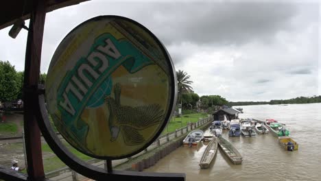 Barcos-Atracados-A-Lo-Largo-De-La-Orilla-Del-Río-Detrás-De-Un-Cartel-De-Cerveza-Aguila-Erosionado