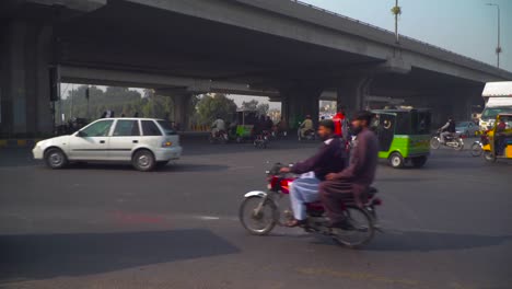 Vueltas-De-Tiempo-De-Tráfico-Y-Personas-Cruzando-La-Calle-En-La-Plaza-Bajo-El-Paso-Elevado,-Una-Ambulancia-Que-Viene,-Rickshaws,-Camiones,-Motocicletas-Que-Se-Detienen-Sobre-El-Paso-De-Cebra,-Luces-De-Las-Calles-Sobre-El-Paso