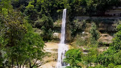 A-beautiful-waterfall-cascades-down-a-cliff-into-a-beautiful-turquoise-pool-below-in-the-middle-of-a-lush-green-jungle-in-Bohol,-Philippines-while-locals-enjoy-the-cool-relaxing-water