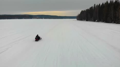 Toma-Aérea-De-Seguimiento-De-Un-Hombre-Conduciendo-Su-Moto-De-Nieve-En-Indalsalven-En-Timra,-Sundsvall,-Suecia