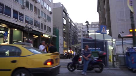 En-La-Cámara-Del-Automóvil-Tomas-Panorámicas-De-Personas,-Automóviles-Y-Tiendas-Por-La-Noche-En-Una-Calle-Muy-Transitada-En-Atenas