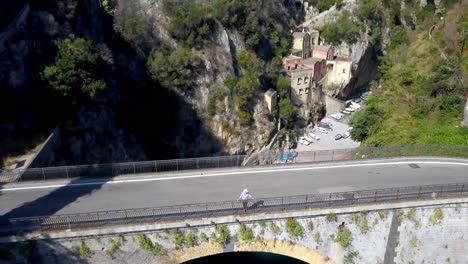 The-arch-bridge-with-man-with-hat-admiring-the-view-while-a-car-passes,-Aerial-pedestal-lift-reveal-shot