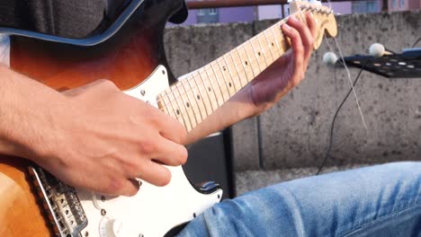 Primer-Plano-De-Un-Guitarrista-Tocando-La-Guitarra-Eléctrica-En-Un-Concierto-Al-Aire-Libre,-Con-Bloques-De-Apartamentos-Coloridos-Visibles-En-El-Fondo