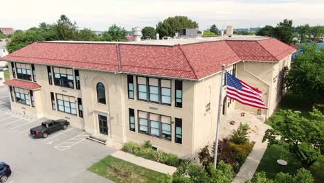 Exterior-Del-Edificio-De-La-Escuela-Pública-Americana,-Bandera-De-Estados-Unidos-Ondeando-En-El-Viento,-Establecimiento-De-Tiro,-Aéreo,-Arquitectura-De-Estilo-Español-Con-Techo-De-Terracota