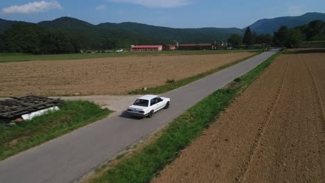 Toma-De-Seguimiento-Aéreo-Sobre-El-Modelo-De-Automóvil-Bmw-E30-Conduciendo-A-Través-De-Campos-Cultivados