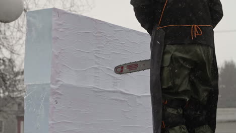 Ice-sculptor-standing-in-front-of-uncut-ice-blocks-with-chainsaw-in-falling-snow,-Slow-Motion