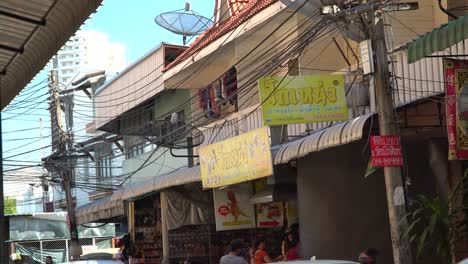 People-are-walking-along-the-street-next-to-the-local-market,-cars-passing-by,-someone-is-eating