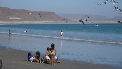 Establecimiento-De-Tiro,-Niños-Jugando-En-La-Playa-De-San-Juanico,-California-Seguro,-México,-Un-Niño-Con-Selfie-Stick-Persiguiendo-A-Los-Pájaros-En-El-Fondo