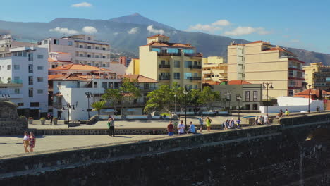 Die-Küstenskyline-Von-Puerto-De-La-Cruz,-Kanarische-Inseln,-Spanien,-Touristen,-Die-An-Einem-Sonnigen-Tag-Auf-Dem-Steinernen-Hafenweg-Spazieren-Und-Die-Aussicht-Genießen,-Im-Hintergrund-Der-Berg-Pico-De-Teide,-4K-Luftaufnahme