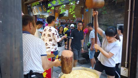 Man-and-woman-hitting-hard-with-big-wooden-hammers-to-crack-grain-which-will-be-used-in-making-of-sweet-snack-for-sale-on-the-street,-China