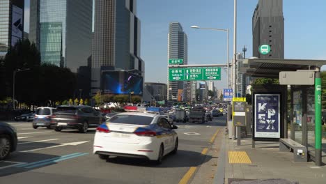 El-Coche-De-Policía-Pasa-Por-Una-Parada-De-Autobús-Vacía-Durante-El-Intenso-Tráfico-En-La-Estación-De-Samseong,-Coex,-Edificio-De-Rascacielos-De-Gran-Altura-De-Seúl-En-El-Fondo-Cuando-Las-Tarjetas-Conducen-Por-La-Carretera-De-Yeongdong