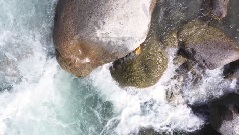 Toma-Aérea-De-La-Corriente-De-Agua-Que-Fluye-Cerca-De-Las-Rocas-Y-La-Montaña-En-El-Fondo