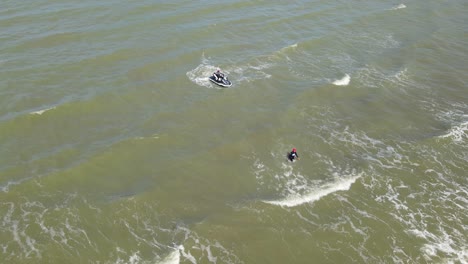 Una-Filmación-Aérea-Descendente-De-Un-Hombre-Vadeando-Hacia-La-Orilla,-Una-Moto-De-Agua-Esperando,-Olas-Corriendo