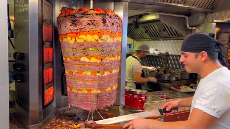 Hombre-Sonriente-Feliz-Cortando-Carne-De-Un-Asador-Vertical-En-Un-Restaurante-De-Kebab-En-Bodrum-Turquía,-Gente-Trabajando-En-La-Cocina,-Buena-Comida-Tradicional,-Carne-Sazonada-Con-Patatas-Y-Zanahorias,-Tiro-De-4k