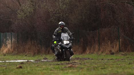 Moto-Salpicando-Estanques-De-Agua-Y-Barro,-Conductor-De-Cámara-Lenta-Lluviosa-Con-Impermeable-Negro-Y-Casco,-Cruz-Extrema-En-Los-Campos