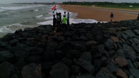 Grupo-De-Hombres-Jóvenes-Patriotas-Sostienen-La-Bandera-Nacional-India-De-Pie-En-Una-Playa-Rocosa-Con-Olas-Rompiendo