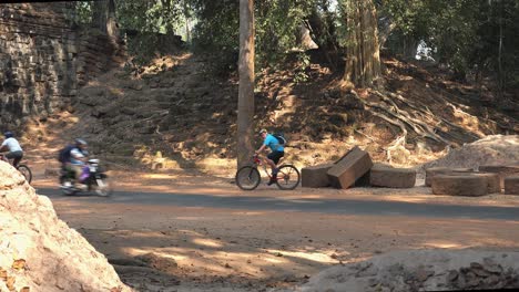 Wide-Exterior-Still-Shot-of-Tourists-on-a-Bicycle-Tour-With-Some-Traffic-on-Road-With-Ancient-Stone-Blocks-and-Trees-and-the-back-ground-in-The-Temple-Area