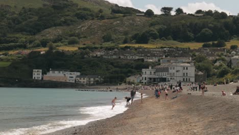 Erwachsene-Und-Kinder-Spielen-An-Einem-Sonnigen-Tag-Am-Strand,-Mit-Häusern-Und-Hügeln-Im-Hintergrund