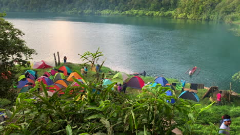 Imágenes-Amplias-De-Personas-Acampando-Junto-Al-Lago-De-Holon,-Con-Una-Carpa-Colorida-Colocada-En-El-Suelo,-Un-Lago-Azul-Y-Un-Bosque-En-El-Fondo