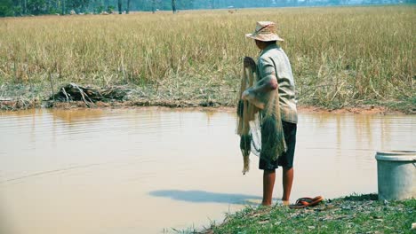 Pescador-Al-Borde-Del-Río-Preparando-Su-Red-Mirando-Alrededor-Para-Seleccionar-El-Lugar-Correcto-Y-Luego-Caminando-Hacia-El-Río