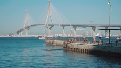 Niño-Salta-Del-Muelle-A-La-Bahía-Con-El-Puente-De-Cádiz-En-El-Fondo,-España,-Cámara-Lenta