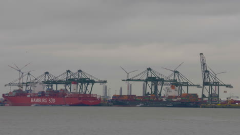 Große-Frachtkähne-Legten-An-Einem-Bewölkten-Tag-Im-Hafen-Von-Antwerpen-An-Und-Be--Und-Entluden-Sendungen-Mit-Windmühlen-In-Der-Ferne-–-Totalaufnahme