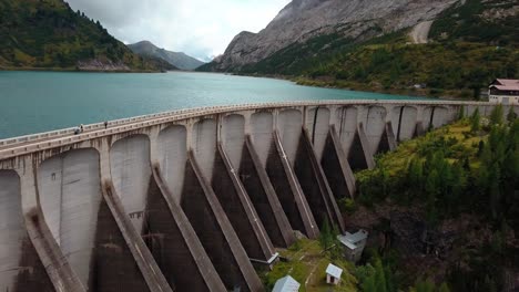 Muro-De-La-Presa-Del-Lago-Fedaia-En-La-Zona-Montañosa-De-Dolomita-En-El-Norte-De-Italia-Con-Gente-Caminando-Sobre-La-Estructura,-Tiro-De-Revelación-De-La-Plataforma-Rodante-De-Drones-Aéreos