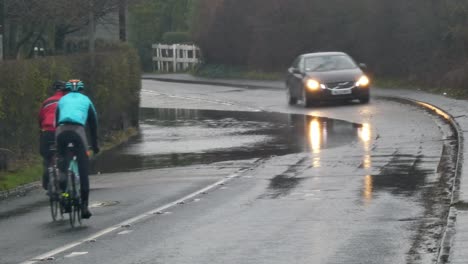 Ciclistas-Andar-En-Bicicleta-Alrededor-De-Stormy-Flash-Inundado-Road-Corner-Bend-Uk