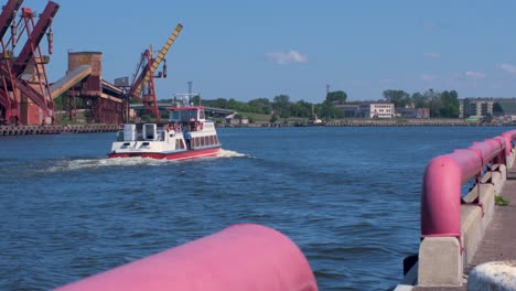 Vergnügungsboot-Hercogs-Jēkabs-Mit-Menschen-Auf-Dem-Deck,-Die-Sich-Am-Fluss-Venta-Bewegen,-Hafenkräne-Und-Hafenlager-Im-Hintergrund,-Sonniger-Sommertag,-Hafen-Von-Ventspils,-Totalaufnahme-Aus-Der-Ferne