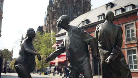 Estatua-De-Bronce-Con-Gente-Y-Piscina-De-Hidromasaje-En-Aquisgrán,-Alemania,-Llamada-Circulación-De-Dinero-Con-La-Catedral-De-Aachener-Dom-En-Segundo-Plano