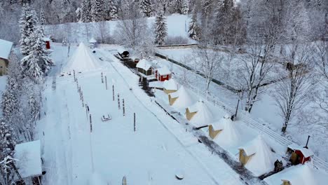 Vista-Aérea-Sobre-árboles-Nevados-Hacia-Cabañas-Y-Tiendas-De-Campaña-En-El-Pueblo-Finlandés-De-Kalevala,-En-Un-Día-De-Invierno-Nublado,-En-Polonia---Aumento,-Disparo-De-Drones