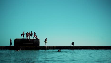 Piscina-De-Agua-Salada-En-La-Costa-Con-Niños-Saltando-Desde-El-Muelle-Sobre-El-Agua-Al-Sol