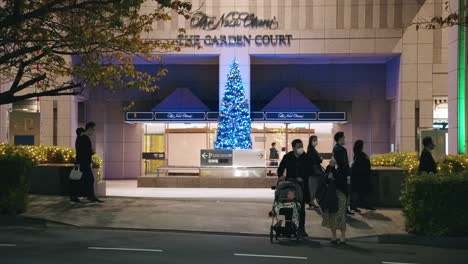 People-Wearing-Masks-In-Front-Of-The-Garden-Court-In-Tokyo-At-Night---Decorative-Christmas-Tree-In-The-Background---slow-motion,-wide-shot