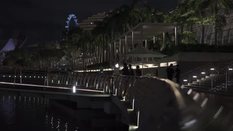 Vista-De-La-Gente-Caminando-Por-El-Paseo-Marítimo-De-Marina-Bay-Sands-Por-La-Noche-Con-El-Volante-De-Singapur-Como-Fondo